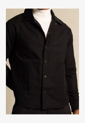 Mann trägt eine schwarze, geknöpfte Jacke über einem weißen Rollkragenpullover vor einem schlichten beigefarbenen Hintergrund.