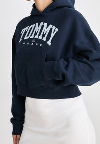 Felpa corta blu navy con logo bianco "TOMMY JEANS", ampio tascone frontale, polsini a costine, materiale in morbido cotone, design minimalista.