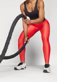 Røde træningsleggins med en glat finish, parret med en sort tanktop. Sort battle rope i hænderne, hvide og sorte sportssko på fødderne.