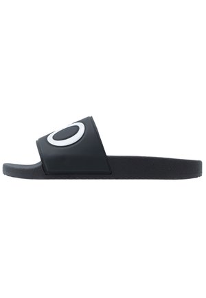 Schwarze Slide-Sandale mit dicker Sohle und breitem Riemen mit weißem ovalem Logo an der Seite, im Seitenprofil vor weißem Hintergrund gezeigt.