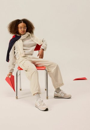 Giovane con capelli ricci e occhiali seduto su una sedia rossa, tiene in mano un aereo di carta rosso, con un altro aereo di carta rosso sul pavimento vicino.