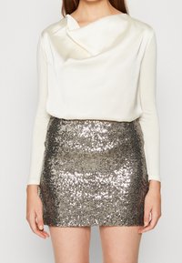 Długo rękawowy kremowy top z opadającym dekoltem, w zestawieniu z krótką spódnicą obszytą srebrnymi cekinami o fakturowej powierzchni.