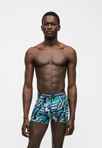 Άνδρας στέκεται μπροστά σε απλό φόντο, φοράει πολύχρωμα μπόξερ με σχέδια και ζώνη Björn Borg, χωρίς πουκάμισο.