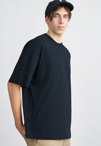 Černé oversized tričko vyrobené z bavlny s kulatým výstřihem, krátkými rukávy a jemným logem na hrudi; hladká textura a pohodlný střih.