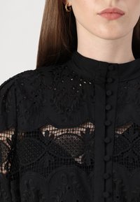 Blusa nera con dettagli in pizzo ricamato, chiusura frontale con bottoni, collo alto e pannelli trasparenti testurizzati.