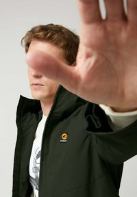 Chaqueta verde oscura con cierre de cremallera, con un logo bordado en el pecho. Textura suave, diseño sin cuello y estilo minimalista.