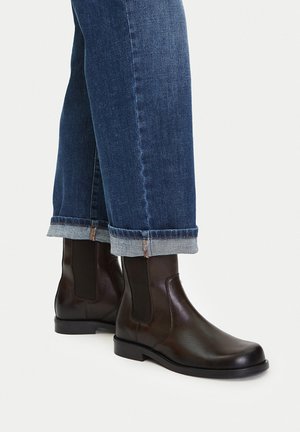 Brune læderankelstøvler med elastiske sidestykker, flade såler og rund tå, parret med opsmøgede mørke denimjeans.