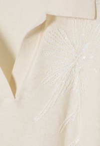 Maglione lavorato a maglia color crema con scollo a V e delicata ricamo di paillettes che ricorda un motivo floreale sul lato. Texture morbida, leggero.