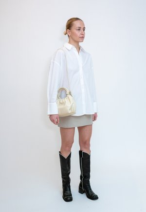 Femme portant une chemise blanche oversize, une mini-jupe beige, des bottes noires montant jusqu'aux genoux et portant un petit sac à main crème sur l'épaule.