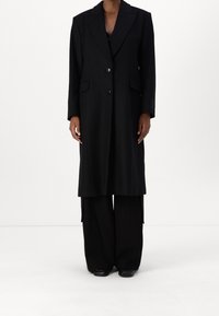 Sort uld langt frakke med notched kl lapel, to knapper, sidelommer og lige silhuet, parret med brede sorte bukser.