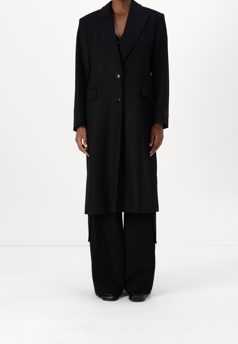 Sort uld langt frakke med notched kl lapel, to knapper, sidelommer og lige silhuet, parret med brede sorte bukser.