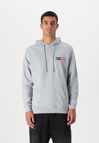 Grå bomulds hoodie med kængurulomme, snøre i hætten og et logo-patch i blå og rød på venstre bryst. Ribstrikkede manchetter og kant.