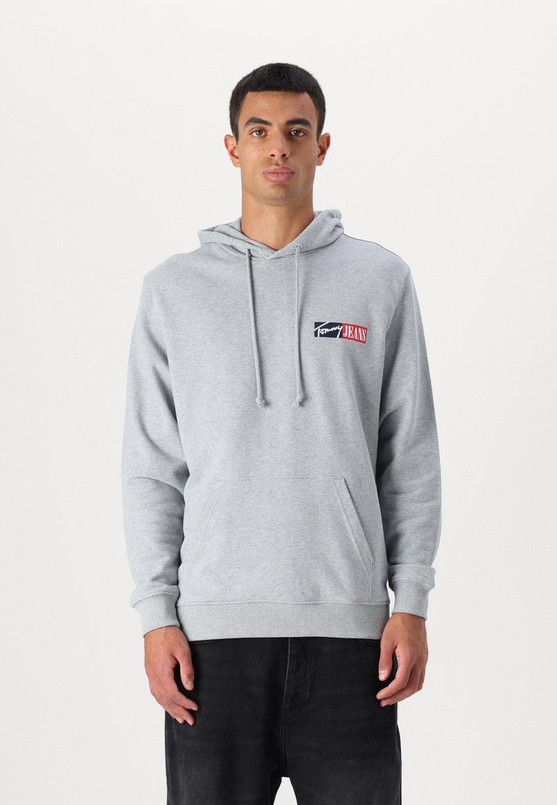 Grå bomulds hoodie med kængurulomme, snøre i hætten og et logo-patch i blå og rød på venstre bryst. Ribstrikkede manchetter og kant.