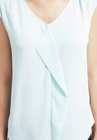 Blusa azzurra chiara con scollatura a V e maniche corte, con un design piegato diagonale lungo la parte anteriore. Tessuto liscio e fluido.