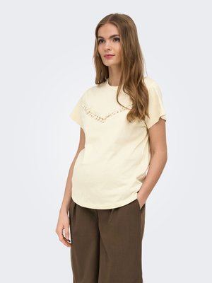 Jonge vrouw met lang bruin haar draagt een crème kortemouwtop met franjes en een losse bruine broek, staand met één hand in de zak.
