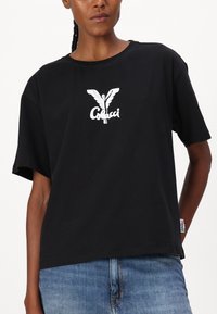 Zwarte katoenen t-shirt met een witte grafiek van een gevleugeld figuur en logo op de borst. Losse pasvorm met korte mouwen en een ronde hals.