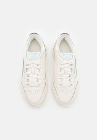 Par av vita Reebok Classic-sneakers med snören, ljusblå logotyp på tungan och perforerade tåhattar, sedda ovanifrån på vit bakgrund.