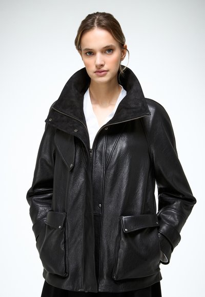 Mujer con chaqueta de cuero negra extragrande con grandes bolsillos y un cuello alto, sobre una camisa blanca, posando frente a un fondo liso.