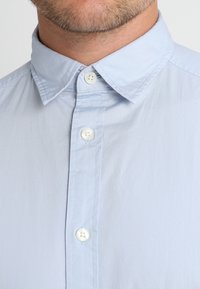 Helleblaue Hemdbluse mit klassischem Kragen, ausgestattet mit weißen Knöpfen und glatter Stoffoberfläche, die die vordere Knopfleiste und Kragendetails zeigt.