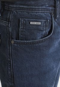Jeans de mezclilla azul oscuro con un bolsillo tipo parche que presenta una etiqueta negra con la inscripción "ANTONY MORATO." El tejido muestra una sutil textura y detalles de costura.
