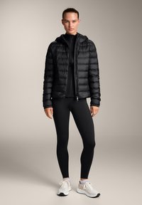 Črna puhovka z vodoravno prešito zasnovo in kapuco, nosena preko črne oprijete majice in črnih pajkic. Bele športne čevlje.