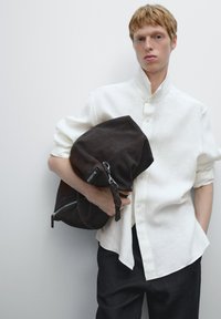 Bruine suede tas met zilveren ritsdetails, vastgehouden tegen een witte linnen blouse met een staande kraag. Donkere broek maakt de look compleet.