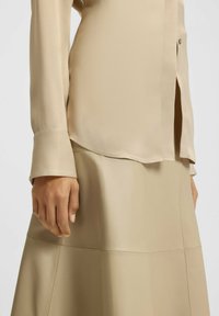 Person, der eine beige Bluse mit langen Ärmeln und einen passenden Lederrock trägt, mit entspanntem Hand an der Seite, vor einem einfarbigen hellen Hintergrund.