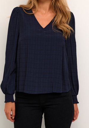 Femme portant un chemisier texturé bleu foncé à manches longues, col en V et poignets smockés, associé à un pantalon noir.