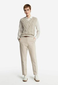 Beżowy sweter bez rękawów z splotem w kablowym wzorze na białej koszuli z kołnierzykiem, w połączeniu z jasnobeżowymi spodniami i białymi sneakersami.