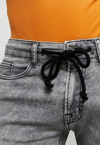 Harmaat farkut, joissa on teksturoitu pinta, mustalla nyörillä vyötäröllä ja tiukka istuvuus. Taskuissa korostusompeleet.