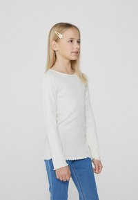 Biała bluzka z długim rękawem i prążkowanym wzorem, z falbaniastym dołem i mankietami, zestawiona z niebieskimi dżinsami; modelka prezentuje profil boczny.