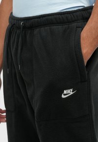 Fekete bolyhos joggerek, elasztikus derékpánttal, húzózsinórral és oldalsó zsebekkel. A bal alsó lábszáron fehér Nike logó található.