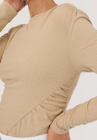 Haut beige à manches longues côtelé présentant un design texturé, une silhouette ajustée et un détail froncé sur le côté, mettant en valeur un tissu doux et subtil.