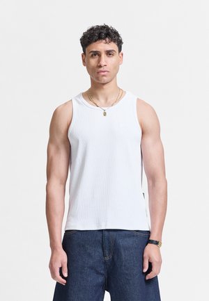 Ung mann som står vendt framover, iført en hvit ermeløs skjorte, mørkeblå jeans, gullkjede og svart armbåndsur.