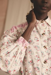 Persoană cu pielea închisă la culoare, purtând o bluză cu model floral, butoni mari, mâneci bufante și ținând o mână aproape de gât.