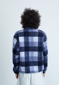 Giacca in fleece con motivo a quadri blu navy e azzurro chiaro, dotata di collo alto e maniche lunghe, con una finitura morbida e texturizzata.