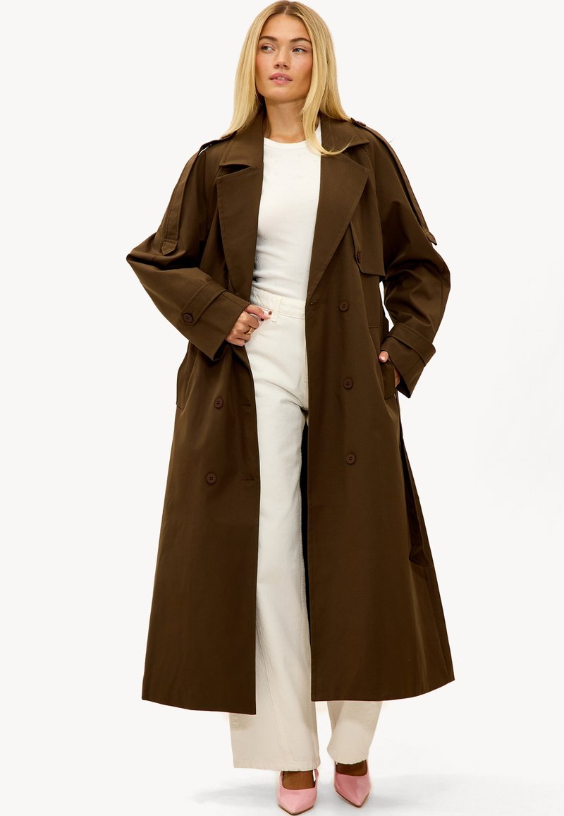 Trench-coat brun à double boutonnage, avec de grands revers et des poches latérales, porté sur un haut blanc et un pantalon clair.