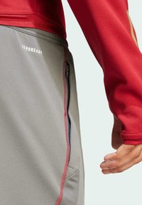 Camiseta roja de manga larga con tejido suave, combinada con pantalones grises que tienen un cierre lateral y detalles de costura roja. Logotipo "AEROREADY" visible.