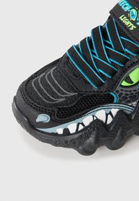 Black sneakers with textured upper, neon blue laces, and a green eye detail. Features white tooth patterns along the sole. Light-up elements included.