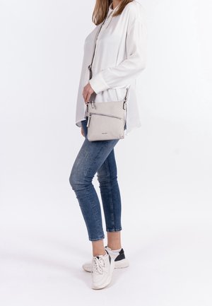 Vrouw draagt een witte blouse, blauwe jeans, witte sneakers en houdt een kleine lichtgrijze crossbodytas met een ritssluiting aan de voorkant vast.