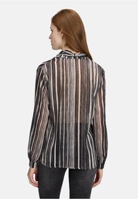 Vrouw met lang rood haar, gekleed in een transparante blouse met verticale strepen in zwart, wit en beige, gecombineerd met donkergrijze broek, gezien van achteren.
