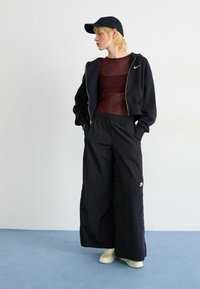 Haut à capuche cropped noir avec fermeture éclair, top ajusté rouge rouille, pantalon large noir et baskets beige. Les accessoires comprennent une casquette noire.