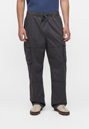 Homme portant un pantalon cargo charbon avec taille à cordon et des baskets beige, debout devant un fond blanc uni.