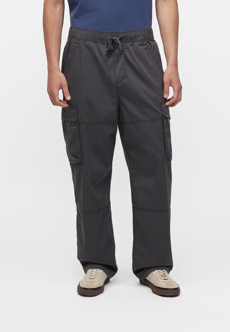 Homme portant un pantalon cargo charbon avec taille à cordon et des baskets beige, debout devant un fond blanc uni.