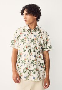 Camicia a maniche corte in tessuto color crema con motivo floreale con foglie verdi, bacche rosse e fiori blu. Design con bottoni sul davanti.