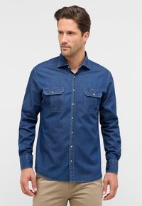Camicia in denim di colore blu scuro, con maniche lunghe, due tasche sul petto e chiusura a bottoni, presentando cuciture a contrasto e un colletto standard.