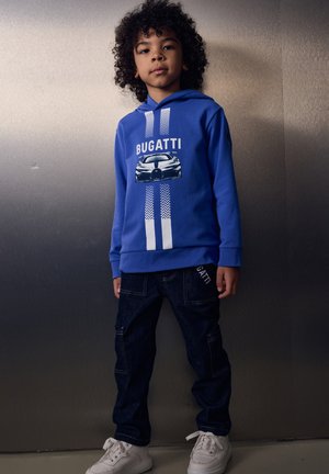 Junge mit lockigem Haar, der einen blauen Bugatti-Hoodie mit einem Autografik, dunkle Jeans und weiße Turnschuhe trägt, steht vor einem schlichten Hintergrund.