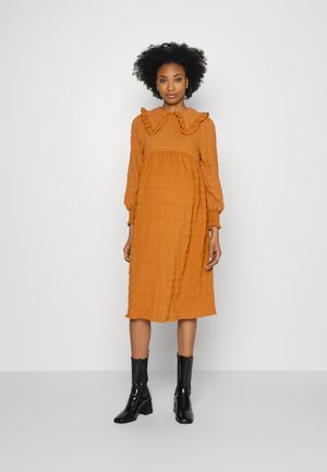 Vestido de mangas compridas, na cor rust, com uma gola texturada eirada e punhos elásticos. Combinado com botas de tornozelo pretas e brilhantes.