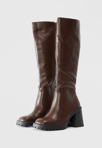 Une paire de bottes hautes en cuir marron avec de gros talons noirs et des fermetures éclair sur le côté, se tenant debout sur un fond blanc uni.