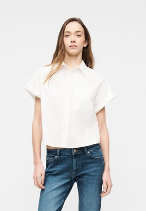 Femme aux longs cheveux bruns portant une chemise blanche à manches courtes boutonnée et un jean bleu, debout devant un fond clair uni.